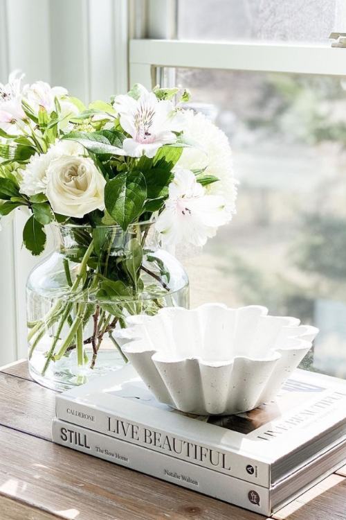 stack of white books on nightstand with fluted bowl and vase of white flowers