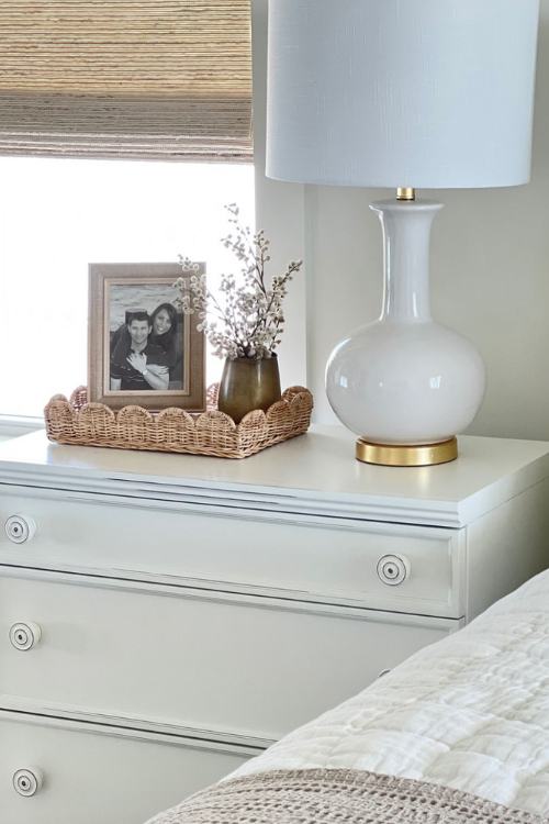 tall white lamp on nightstand with picture frame and vase of dried flowers