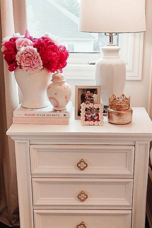 white nightstand with stacked pink books, white lamp and vase of pink flowers